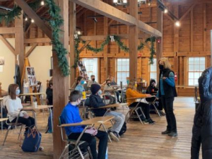 A group of students sit at desks while an instructor teaches before them.