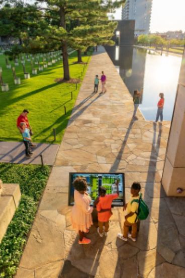 Several families and individual people walk through a serene outdoor space with a stone walkway and reflecting pool nearby.